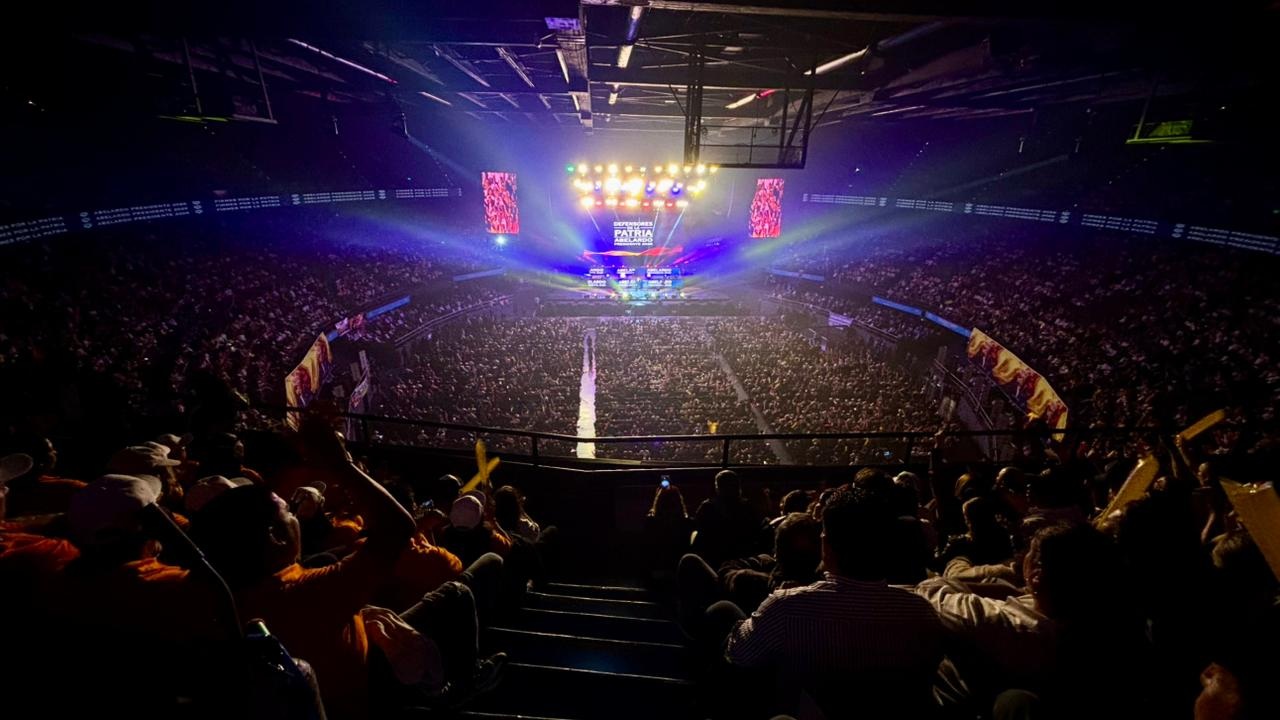 Panorámica del Movistar Arena lleno durante la convención “Firmes por la Patria”. El público ondea banderas de Colombia mientras se observa el escenario iluminado y la pantalla central con mensajes del evento.